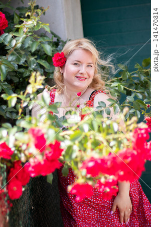 Adult blonde woman smiles with a red rose tucked into her hair, framed by softly blurred blossoms and leafy branches outdoors. Adult blonde woman smiles with a red rose tucked into her hair, framed by softly blurred blossoms and leafy branches outdoors. 141470814