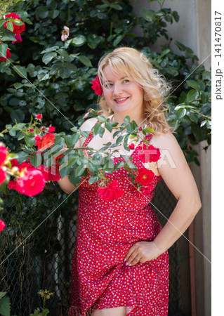 Adult blonde woman smiles at the camera while holding a blooming rose branch and lightly lifting the hem of her red floral dress. 141470817