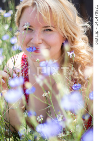 Close portrait of an adult woman smiling at the camera while holding blue cornflower stems among softly blurred summer wildflowers. 141470821