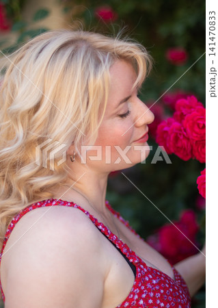 Tight vertical portrait of an adult blonde woman with closed eyes enjoying red rose fragrance against a softly blurred garden. Tight vertical portrait of an adult blonde woman with closed eyes enjoying red rose fragrance against a softly blurred garden. 141470833