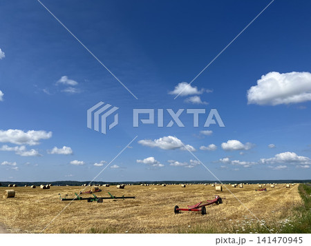 Farm trailers in harvested field with round hay bales across rural landscape under blue summer sky Farm trailers in harvested field with round hay bales across rural landscape under blue summer sky 141470945