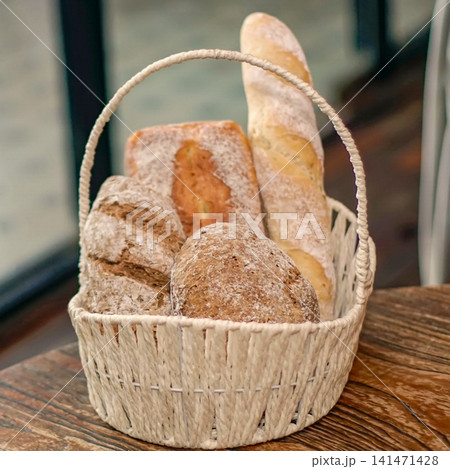 Assortment of Fresh Artisan Breads in a Rustic Woven Basket, Including Baguette and Whole Grain Loaves, Presented on a Wooden Table in a Cozy Bakery Setting 141471428