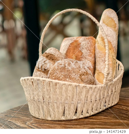 Assortment of Fresh Artisan Breads in a Rustic Woven Basket, Including Baguette and Whole Grain Loaves, Presented on a Wooden Table in a Cozy Bakery Setting 141471429