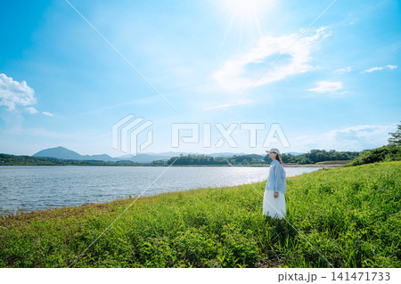 湖畔の草原に立つ白ワンピースの女性と青空 141471733