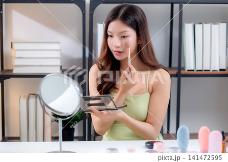 Happy young asian woman applying makeup with a brush on cheek looking mirror. Happy young asian woman applying makeup with a brush on cheek looking mirror. 141472595
