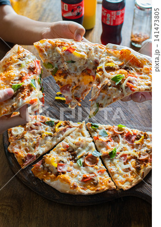 People sharing freshly baked deluxe pizza with melted cheese and assorted toppings on rustic wooden serving board, casual restaurant dining, friendship, family meal and Italian food concept People sharing freshly baked deluxe pizza with melted cheese and assorted toppings on rustic wooden serving board, casual restaurant dining, friendship, family meal and Italian food concept 141473805