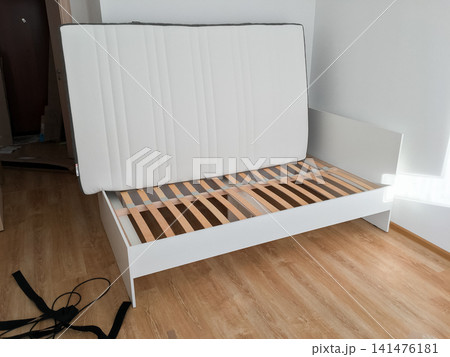 Assembled white bed frame with wooden slat base in an empty room with laminate floor. Assembled white bed frame with wooden slat base in an empty room with laminate floor. 141476181