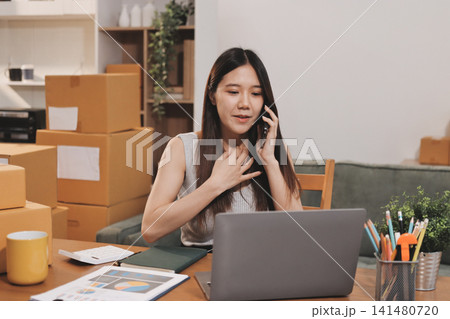 Startup SME small business entrepreneur of freelance Asian woman wearing apron using laptop and box to receive and review orders online to prepare to pack sell to customers, online sme business ideas. Startup SME small business entrepreneur of freelance Asian woman wearing apron using laptop and box to receive and review orders online to prepare to pack sell to customers, online sme business ideas. 141480720