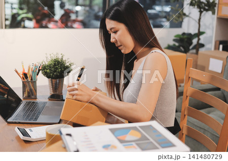 Startup SME small business entrepreneur of freelance Asian woman wearing apron using laptop and box to receive and review orders online to prepare to pack sell to customers, online sme business ideas. Startup SME small business entrepreneur of freelance Asian woman wearing apron using laptop and box to receive and review orders online to prepare to pack sell to customers, online sme business ideas. 141480729