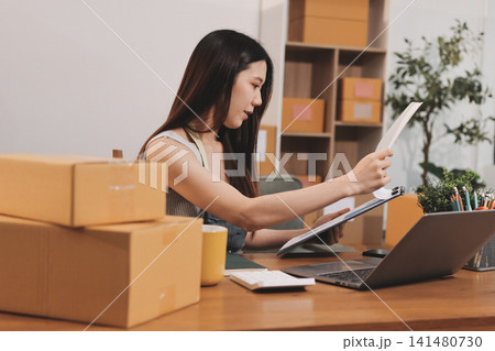 Startup SME small business entrepreneur of freelance Asian woman wearing apron using laptop and box to receive and review orders online to prepare to pack sell to customers, online sme business ideas. Startup SME small business entrepreneur of freelance Asian woman wearing apron using laptop and box to receive and review orders online to prepare to pack sell to customers, online sme business ideas. 141480730