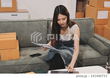 Startup SME small business entrepreneur of freelance Asian woman wearing apron using laptop and box to receive and review orders online to prepare to pack sell to customers, online sme business ideas. Startup SME small business entrepreneur of freelance Asian woman wearing apron using laptop and box to receive and review orders online to prepare to pack sell to customers, online sme business ideas. 141480733