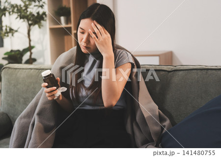 sick asian young woman sneezing into tissue paper while covered with a blanket and sitting on sofa in the living room sick asian young woman sneezing into tissue paper while covered with a blanket and sitting on sofa in the living room 141480755