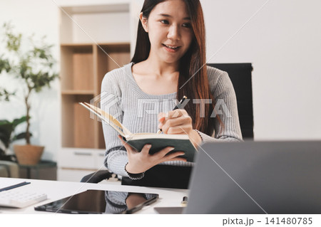 Young asian businesswoman is smiling and working on a laptop while sitting at her desk in a modern office Young asian businesswoman is smiling and working on a laptop while sitting at her desk in a modern office 141480785