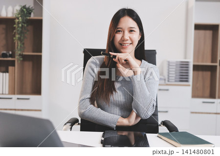 young woman is working on laptop in modern office environment, showcasing focused and productive atmosphere. She is dressed in professional white shirt, with coffee cup and small plant on table, 141480801