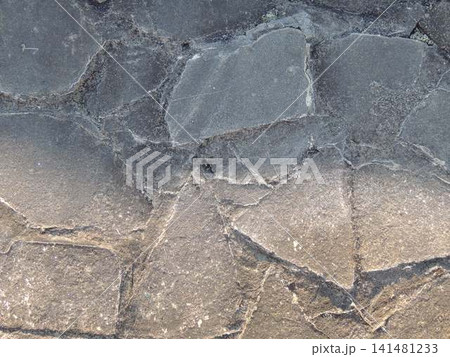 A close-up of an old pavement made of flat, irregularly shaped gray stones is shown. The large elements are bonded with cement mortar, and cracks, chips, and a rough texture are visible on the surface 141481233
