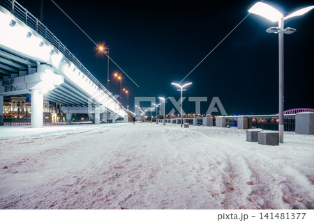 City winter embankment with lanterns at night. Part of the overpass of the bridge and the lanterns on it are visible. Snow on the path stretching into the distance.Ukraine. Kyiv 141481377