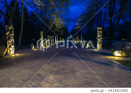 The tiled road in the night park with stone idols illuminated by lamps. Stone statues in the park at night. Illumination of a park road with lanterns at night. Lutsk The tiled road in the night park with stone idols illuminated by lamps. Stone statues in the park at night. Illumination of a park road with lanterns at night. Lutsk 141481400