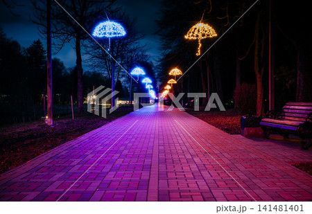The tiled road in the night park with illuminated umbrellas. Benches in the park at night. Illumination of a park road with small lanterns on umbrellas suspended above the path at night. Lutsk.Ukraine The tiled road in the night park with illuminated umbrellas. Benches in the park at night. Illumination of a park road with small lanterns on umbrellas suspended above the path at night. Lutsk.Ukraine 141481401