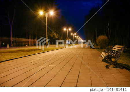 Night city park. Wooden benches, street lights and park alley. The tiled road in the night park with lanterns. Benches in the park at night. Illumination of a park road with lanterns at night. Lutsk Night city park. Wooden benches, street lights and park alley. The tiled road in the night park with lanterns. Benches in the park at night. Illumination of a park road with lanterns at night. Lutsk 141481411