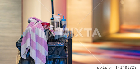 A janitorial trolley loaded with cleaning equipment stands in a hotel corridor. The blurry background shows a carpeted hallway with warm lighting. A janitorial trolley loaded with cleaning equipment stands in a hotel corridor. The blurry background shows a carpeted hallway with warm lighting. 141481828