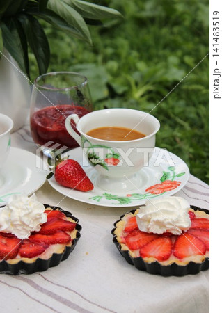 Fresh strawberry tarts with whipped cream served alongside coffee in a decorative cup and a glass of raspberry drink on a picnic blanket in a garden setting Fresh strawberry tarts with whipped cream served alongside coffee in a decorative cup and a glass of raspberry drink on a picnic blanket in a garden setting 141483519