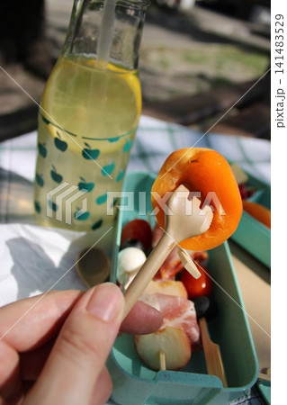 Hand holding a small fork with an apricot slice above a colorful picnic box filled with assorted fruits and a bottle of lemonade on a checkered tablecloth 141483529