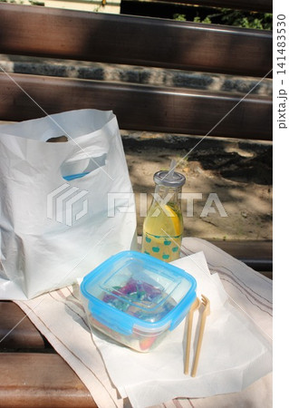 Lunch picnic setup featuring a white takeout bag, a clear beverage bottle with lemon slices, and a blue container with colorful food on a wooden bench Lunch picnic setup featuring a white takeout bag, a clear beverage bottle with lemon slices, and a blue container with colorful food on a wooden bench 141483530