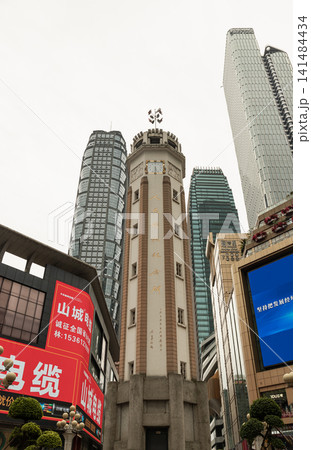 A historic octagonal clock tower stands in the center of a modern city center. Tall skyscrapers and large commercial buildings with digital screens surround the monument under an overcast sky. A historic octagonal clock tower stands in the center of a modern city center. Tall skyscrapers and large commercial buildings with digital screens surround the monument under an overcast sky. 141484434
