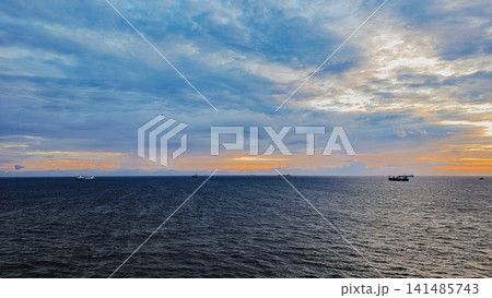 Wide angle view of commercial cargo ships floating on sea horizon under colorful sunset sky for global maritime logistics shipping and travel background Wide angle view of commercial cargo ships floating on sea horizon under colorful sunset sky for global maritime logistics shipping and travel background 141485743