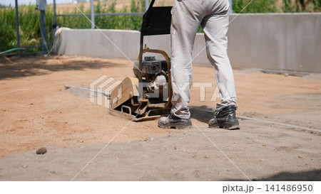 Worker in work pants and safety boots uses plate compactor to flatten soil near concrete wall and metal framing, preparing residential ground for foundation work under clear Worker in work pants and safety boots uses plate compactor to flatten soil near concrete wall and metal framing, preparing residential ground for foundation work under clear 141486950