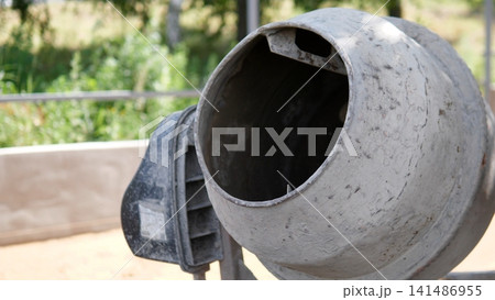 Weathered concrete mixer drum in close up on outdoor construction site, with worn metal surface, open circular mouth, blurred green background, natural daylight, industrial Weathered concrete mixer drum in close up on outdoor construction site, with worn metal surface, open circular mouth, blurred green background, natural daylight, industrial 141486955