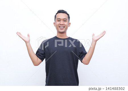 Young man smiles warmly with open arms. He wears a solid black t-shirt and stands against plain white background. His cheerful expression conveys openness and friendliness Young man smiles warmly with open arms. He wears a solid black t-shirt and stands against plain white background. His cheerful expression conveys openness and friendliness 141488302