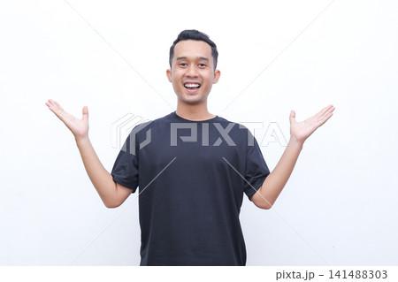 Young man smiles warmly with open arms. He wears a solid black t-shirt and stands against plain white background. His cheerful expression conveys openness and friendliness Young man smiles warmly with open arms. He wears a solid black t-shirt and stands against plain white background. His cheerful expression conveys openness and friendliness 141488303