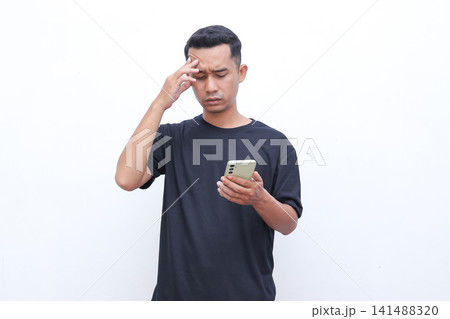 Man holds phone, eyes closed, hand to forehead. Looks stressed or confused, possibly scrolling. Simple white background shows clear focus. Ideal for digital stress, work pressure, or decision fatigue Man holds phone, eyes closed, hand to forehead. Looks stressed or confused, possibly scrolling. Simple white background shows clear focus. Ideal for digital stress, work pressure, or decision fatigue 141488320