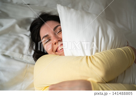 Smiling woman embracing a soft pillow in a cozy bedroom setting. Restful sleep comfort relaxation and bedding advertisement 141488805