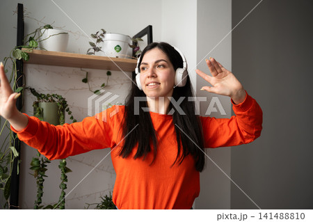Smiling young woman wearing headphones gesturing in home office setting. Remote work communication and online presentation Smiling young woman wearing headphones gesturing in home office setting. Remote work communication and online presentation 141488810
