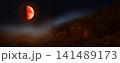 Blood moon with orange surface over forest at night. Half of the moon is illuminated. 141489173