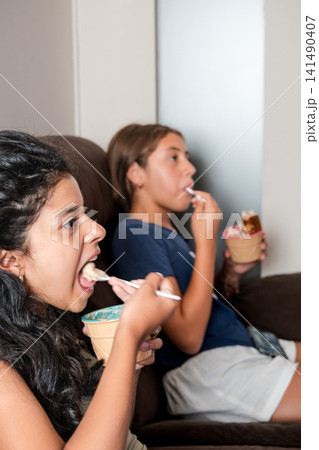 Two hispanic girls eating ice cream on sofa watching tv Two hispanic girls eating ice cream on sofa watching tv 141490407