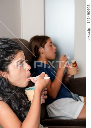 Two hispanic girls eating ice cream on sofa watching tv Two hispanic girls eating ice cream on sofa watching tv 141490408