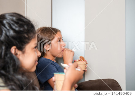 Two hispanic girls eating ice cream on sofa watching tv 141490409