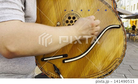 Male fingers of musician playing the bandura on a sunny summer day. Sunlight falls on the strings and wood surface, creating a warm and lively scene of outdoor music and authentic street performance 141490515