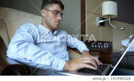 Handsome entrepreneur use notebook for work sitting in armchair at home interior. Young businessman in glasses working on laptop in modern hotel room. Manager showing remote job and productivity 141490526