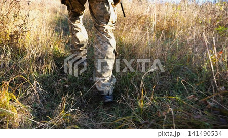 Low view to male feet of ukrainian soldiers going along trail at countryside. Legs of young army man walking through dry grass at sunny day. War in Ukraine. Slow motion Low view to male feet of ukrainian soldiers going along trail at countryside. Legs of young army man walking through dry grass at sunny day. War in Ukraine. Slow motion 141490534