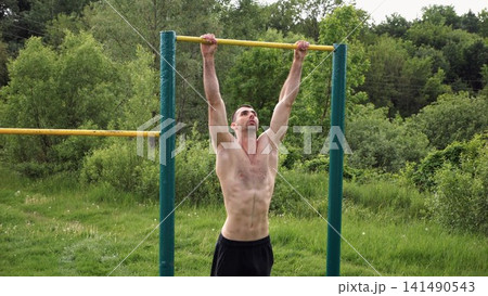 Muscular athlete doing pull ups on horizontal bar at sports ground. Young shirtless sportsman exercising at nature. Strong sporty man training outdoor. Concept of healthy and active lifestyle. Slow mo Muscular athlete doing pull ups on horizontal bar at sports ground. Young shirtless sportsman exercising at nature. Strong sporty man training outdoor. Concept of healthy and active lifestyle. Slow mo 141490543