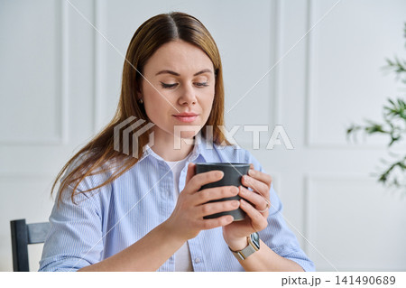 Pensive woman in blue shirt holding cup and looking away Pensive woman in blue shirt holding cup and looking away 141490689