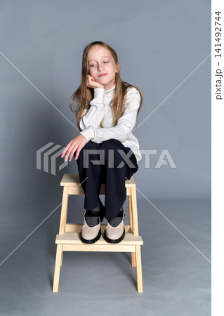 Girl sits wooden step stool with hands gently placed on her knee, subtle smile and quiet confidence Girl sits wooden step stool with hands gently placed on her knee, subtle smile and quiet confidence 141492744