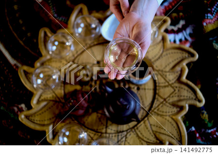 Top view of hands presenting a clear teacup over a floral tea tray with cups and teapot during a quiet indoor ritual. Tea meditation and slow routines help reduce stress and restore emotional balance. Top view of hands presenting a clear teacup over a floral tea tray with cups and teapot during a quiet indoor ritual. Tea meditation and slow routines help reduce stress and restore emotional balance. 141492775