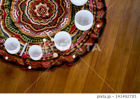 Top view of white crystal singing bowls, soft mallets, and small candles arranged on a colorful rug with wooden copy space. Sound meditation and slow rituals support calm, focus, and stress relief. 141492785