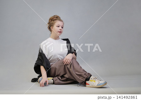 Seated and stretching her arms, a woman in casual attire including a white t-shirt, black denim jacket, and beige pants poses thoughtfully against a simple background. This photo captures a relaxed Seated and stretching her arms, a woman in casual attire including a white t-shirt, black denim jacket, and beige pants poses thoughtfully against a simple background. This photo captures a relaxed 141492861