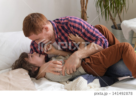 Delighted father and kid connecting through lighthearted tickling, generating a cozy, cheerful ambiance in their bedroom. importance of parent-child bonding and its effects on child development. Delighted father and kid connecting through lighthearted tickling, generating a cozy, cheerful ambiance in their bedroom. importance of parent-child bonding and its effects on child development. 141492878